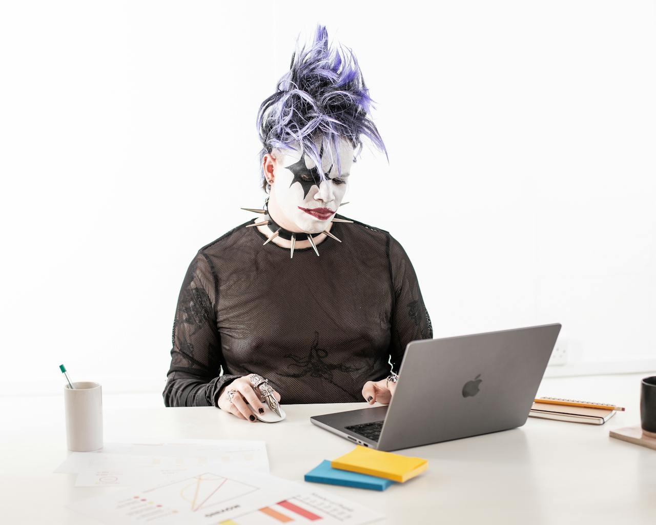 A goth-styled man with purple hair works on a laptop in a bright studio setting, showcasing modern Halloween office vibes.